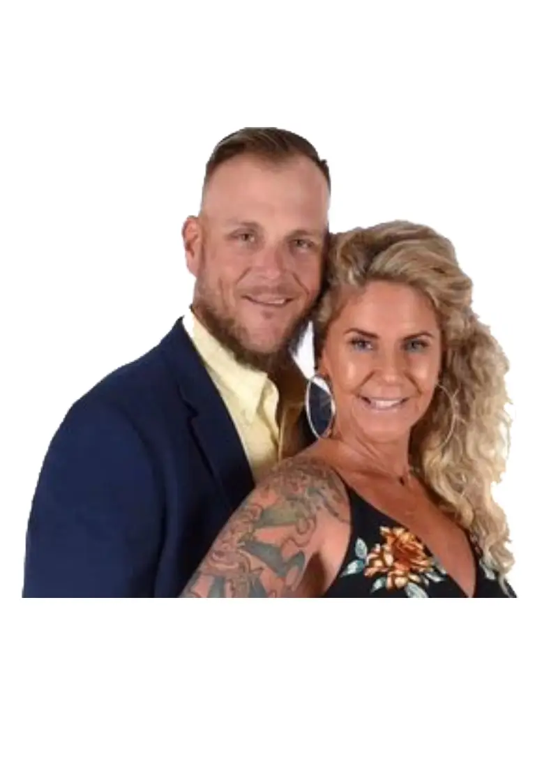 A man in a dark suit and a woman in a floral dress pose closely together, showcasing the woman’s intricate tattoos.